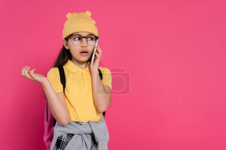 schoolgirl in beanie hat and glasses talking smartphone on pink background, stylish look, phone call
