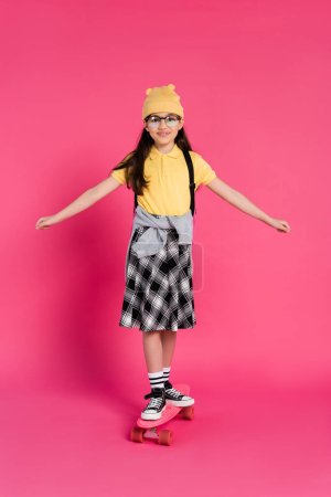 glücklich Schulmädchen mit Hut und Brille Reiten Penny Board auf rosa Hintergrund, stilvollen Look