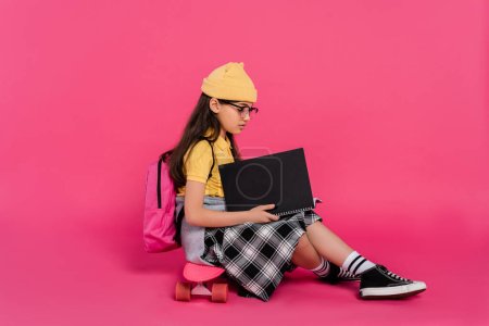 stylish schoolgirl in beanie hat and glasses sitting on penny board, pink background, notebooks