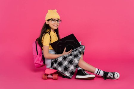 happy schoolgirl in beanie hat and glasses sitting on penny board, pink background, notebooks