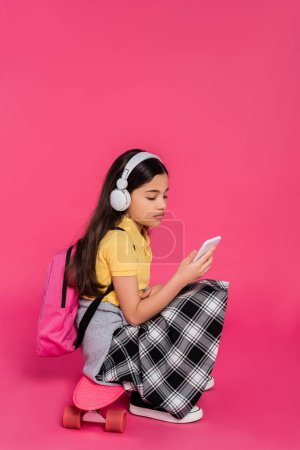 écolière dans des écouteurs sans fil assis sur une planche de penny, fond rose, en utilisant un smartphone