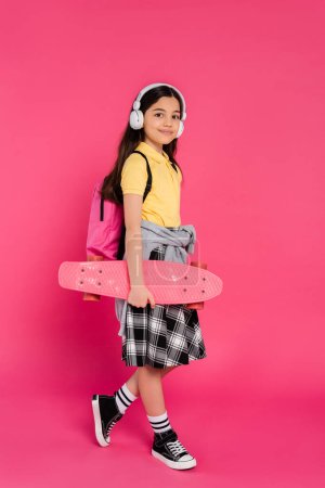 Foto de Colegiala feliz en auriculares inalámbricos de pie con penny board, fondo rosa, después de clases - Imagen libre de derechos
