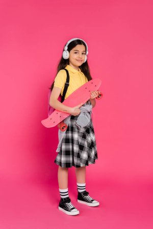 Foto de Alegre chica en auriculares inalámbricos de pie con penny board, fondo rosa, después de clases - Imagen libre de derechos