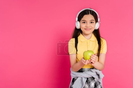 happy schoolgirl in wireless headphones holding green apple on pink background, kid with backpack