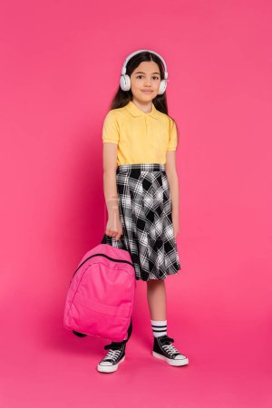 smiling schoolgirl in wireless headphones holding backpack on pink background, brunette student