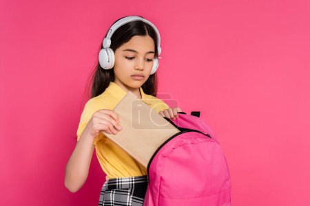 brünettes Schulmädchen mit drahtlosen Kopfhörern, das Buch in den Rucksack steckt, rosa Hintergrund, Studentin