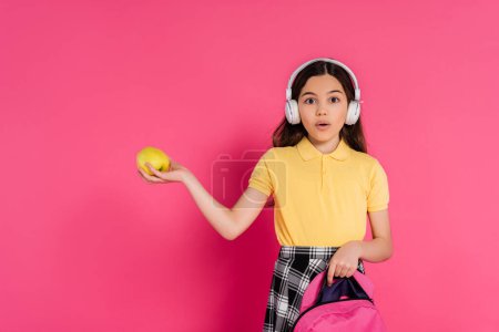 Schockierte Schülerin in drahtlosen Kopfhörern mit Apfel und Rucksack, rosa Hintergrund, Studentin