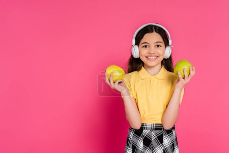 surprised schoolgirl in wireless headphones holding apple and backpack, pink background, student
