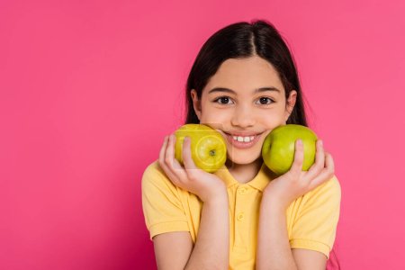 positive girl with brunette hair holding green apples on pink background, portrait, happiness