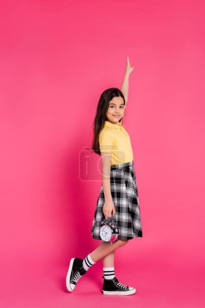 Photo for Happy schoolgirl holding alarm clock on pink background, looking at camera, pointing up, full length - Royalty Free Image