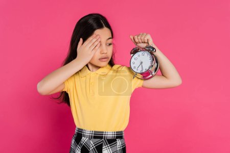 brunette schoolgirl looking at vintage alarm clock isolated on pink, back to school concept,