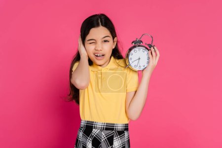 schoolgirl looking at camera, winking, holding vintage alarm clock isolated on pink, back to school