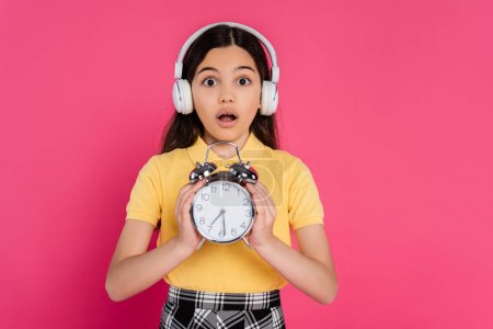 shocked schoolgirl in wireless headphones holding vintage alarm clock isolated on pink, student life