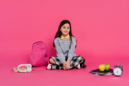 happy schoolgirl sitting near backpack, notebooks, headphones, green apples, vintage alarm clock
