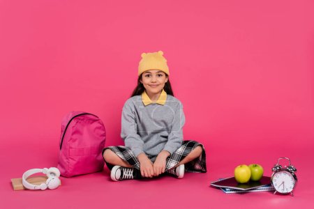 happy schoolgirl in beanie hat sitting near backpack, notebooks, headphones, apples and alarm clock