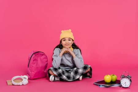 glückliches Schulmädchen mit Mütze, das neben Notizbüchern, Kopfhörern, Äpfeln, Rucksack und Wecker sitzt