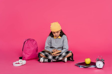 schoolgirl in beanie hat reading book, sitting near headphones, apple, backpack, alarm clock