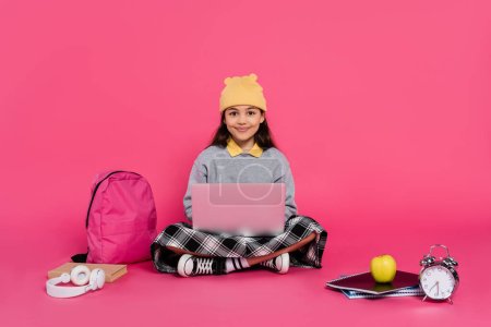 colegiala feliz en sombrero de gorro usando el ordenador portátil, sentado cerca de los auriculares, manzana, mochila, despertador