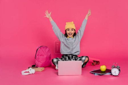 Foto de Alegre chica en gorro sombrero usando el ordenador portátil, sentado cerca de los auriculares, manzana, mochila, despertador - Imagen libre de derechos