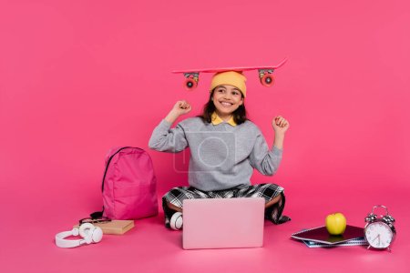 Photo for Happy girl in beanie hat sitting with penny board on head, laptop, headphones, apple,  alarm clock - Royalty Free Image