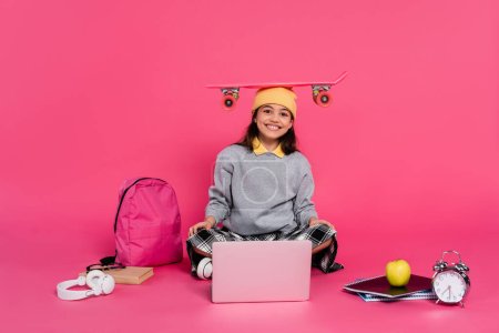 sonrisa, chica en gorro sombrero sentado con penny board en la cabeza, ordenador portátil, auriculares, manzana, reloj despertador