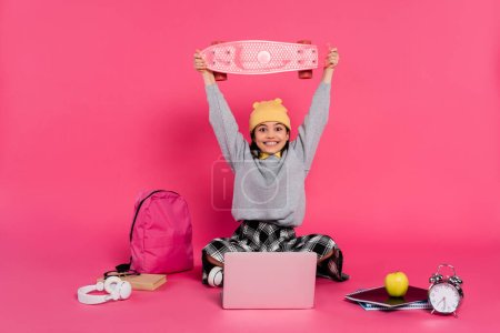 Photo for Excited, girl in beanie hat holding penny board on head, laptop, headphones, apple,  alarm clock - Royalty Free Image