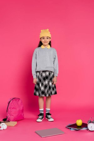 happy schoolgirl in beanie hat standing on pink, laptop, headphones, apple, alarm clock, notebooks