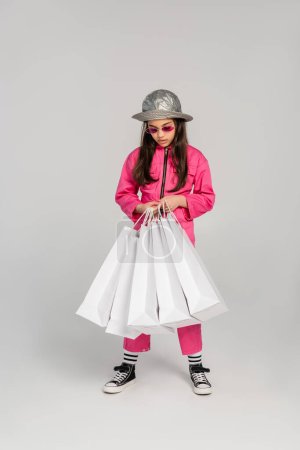 elegante chica en traje rosa y sombrero de panama sosteniendo bolsas de compras sobre fondo gris, longitud completa