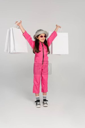 aufgeregtes Mädchen in stylischem rosa Outfit und Panamahut mit Einkaufstaschen auf grauem Hintergrund