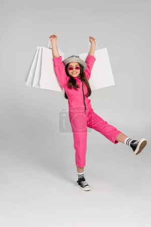 fröhliches Mädchen in stylischem rosa Outfit und Panamahut mit Einkaufstaschen auf grauem Hintergrund