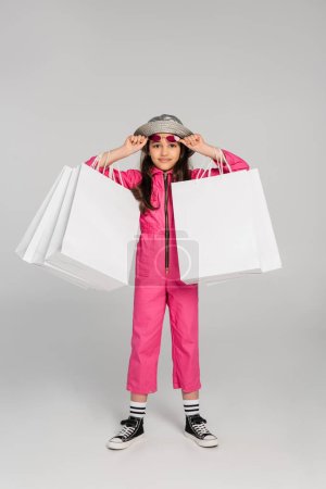girl in stylish outfit and panama hat holding shopping bags on grey, adjusting pink sunglasses