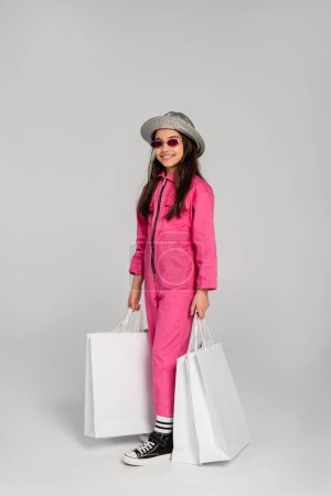 chica alegre en traje elegante, gafas de sol y sombrero panama sosteniendo bolsas de compras sobre fondo gris