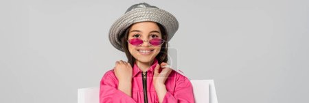 Foto de Chica positiva en traje elegante, gafas de sol y sombrero de panama posando con bolsas de compras en gris, bandera - Imagen libre de derechos