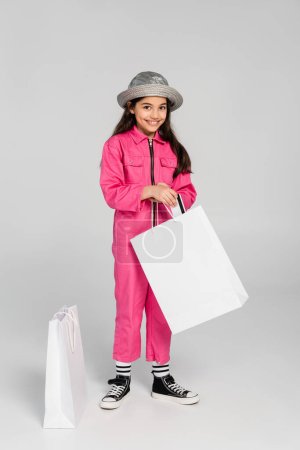 happy girl in trendy outfit and panama hat putting credit card into shopping bags, grey background