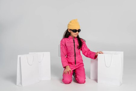 modisches Mädchen mit Beanie-Hut und Sonnenbrille sitzt neben Einkaufstüten auf grauem, glücklichem Kind, trendy