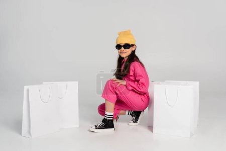 Foto de Chica de moda en gorro sombrero y gafas de sol sentado posando cerca de bolsas de compras en gris, niño feliz - Imagen libre de derechos