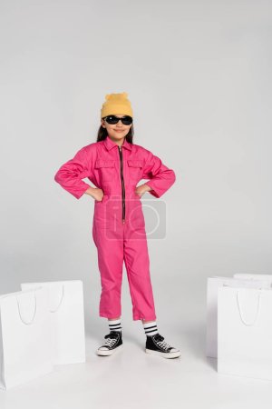 modisches Mädchen mit Mütze und Sonnenbrille, das mit Händen auf Hüften neben Einkaufstaschen posiert