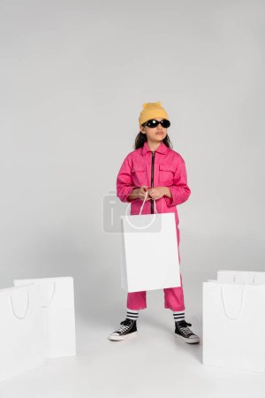 Stylisches Preteen Girl mit Beanie-Hut und Sonnenbrille steht und hält Einkaufstüten auf grau