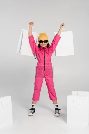aufgeregtes Mädchen mit Mütze und Sonnenbrille mit Einkaufstaschen auf grauem Hintergrund, Stil