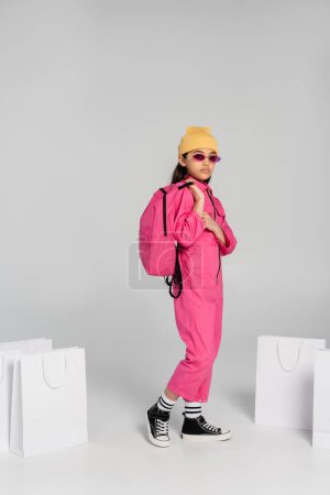 Foto de De vuelta a la escuela, chica con estilo en gorro y gafas de sol con bolsas de compras y mochila en gris - Imagen libre de derechos