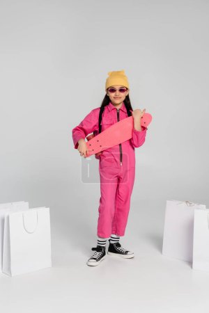 Foto de Chica feliz y elegante en gorro y gafas de sol sosteniendo penny board cerca de bolsas de compras en gris - Imagen libre de derechos