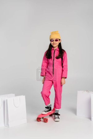 happy girl in beanie and stylish sunglasses riding penny board, shopping bags on grey background