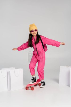 cheerful girl in beanie and stylish sunglasses riding penny board, shopping bags on grey background