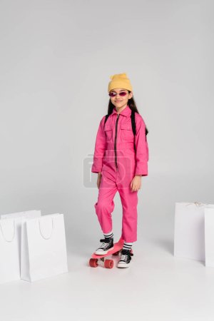 Positives Mädchen in Mütze und rosa Sonnenbrille auf Penny Board, Einkaufstaschen auf grauem Hintergrund