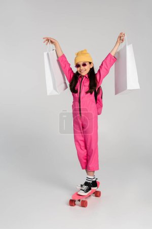 happy girl in beanie and pink sunglasses riding penny board and holding shopping bags on grey