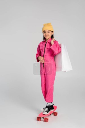 glückliches Mädchen mit Mütze und rosa Outfit reitet Penny Board und hält Einkaufstüten auf grau