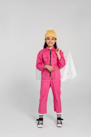 Foto de Chica feliz en gorro amarillo sombrero y traje rosa sosteniendo bolsas de compras y de pie en gris, moda - Imagen libre de derechos