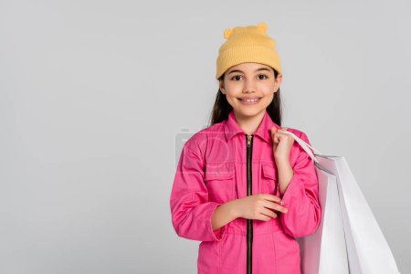 fröhliches Mädchen mit Mütze und rosa Outfit, Einkaufstaschen in der Hand und nach oben zeigend, grauer Hintergrund