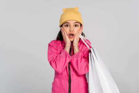 amazed girl in beanie hat and pink outfit holding shopping bags and looking at camera, grey backdrop