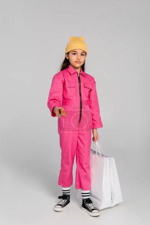stylisches Mädchen mit Mütze, Kreditkarte und Einkaufstaschen auf grauem, trendigem Outfit, Kindereinkauf